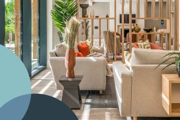 A modern living room with beige sofas, wooden partition, plants, and large windows allowing natural light. Decorative cushions and a vase are on the coffee table. Geometric shapes overlay the image.