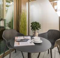 A small outdoor balcony with two black chairs and a round table. On the table are a white cup and saucer, two books, and a plant in a silver pot. The setup overlooks a building.