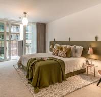 Modern bedroom with a large bed, green headboard, and neatly arranged pillows. There is a side table with lamps, a desk on the left, and a balcony accessible through sliding glass doors in the background.