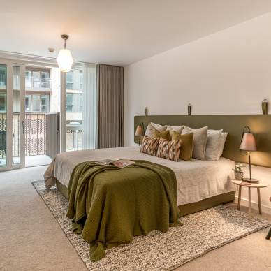 A modern bedroom with a large bed, green and white decor, a desk, chairs, and a window opening to a balcony.