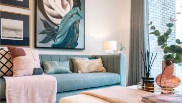 A modern living room featuring a teal sofa with cushions and a beige throw, a wall with abstract artwork, a lit table lamp, and a coffee table with decor items including a pink vase and reed diffuser.