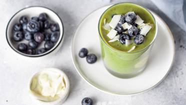 Blueberry, chia seeds, kale and coconut smoothie A glass of green smoothie topped with blueberries and coconut flakes on a white plate, with bowls of blueberries and coconut flakes nearby.