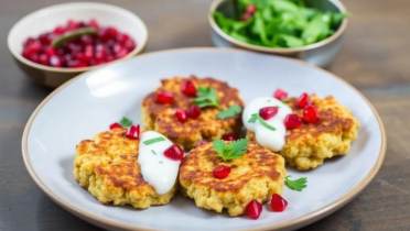 Pumpkin seeds chickpea cauliflower fritters with a pomegranate, mint and coriander soya yoghurt relish Three golden fritters topped with white sauce, pomegranate seeds, and parsley are served on a plate, with bowls of salad and pomegranate seeds in the background.